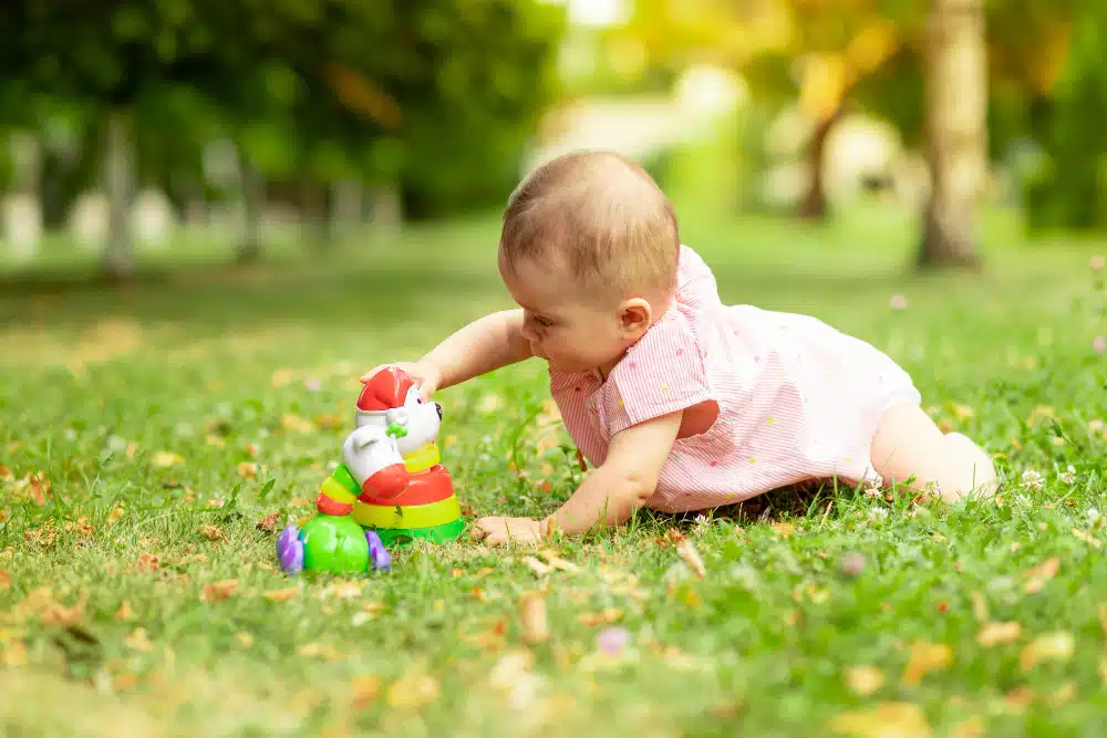 Um bebê engatinhando para alcançar um brinquedo na grama, um brinquedo adequado para o desenvolvimento dos bebês.