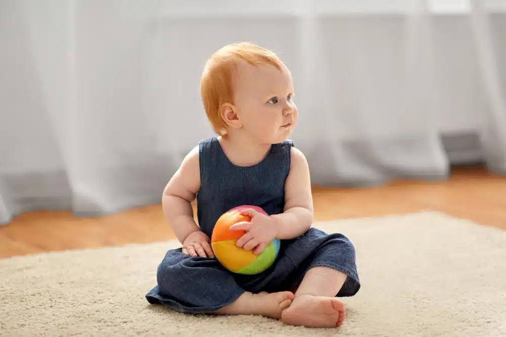 Um bebê sentado num tapete segurando uma bola de pano colorida, um brinquedo Montessori.